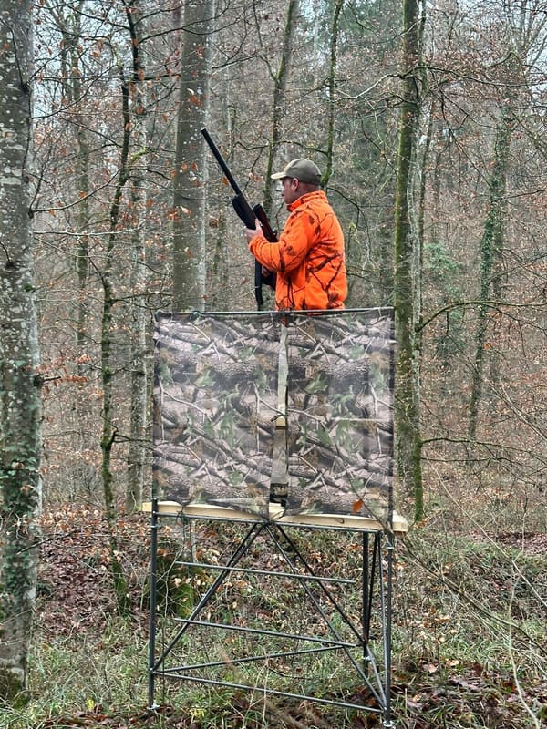 Jäger mit Gewehr auf dem Drückjagdbock mit Tarnnetz im Laubwald